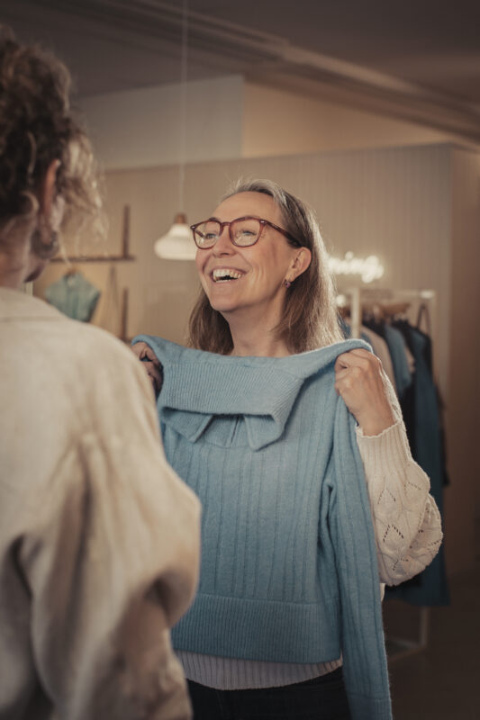 Leende kvinna visar upp blå tröja för annan kvinna i klädbutik
